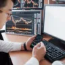 Over the shoulder view of and stock broker trading online while accepting orders by phone. Multiple computer screens ful of charts and data analyses in background.