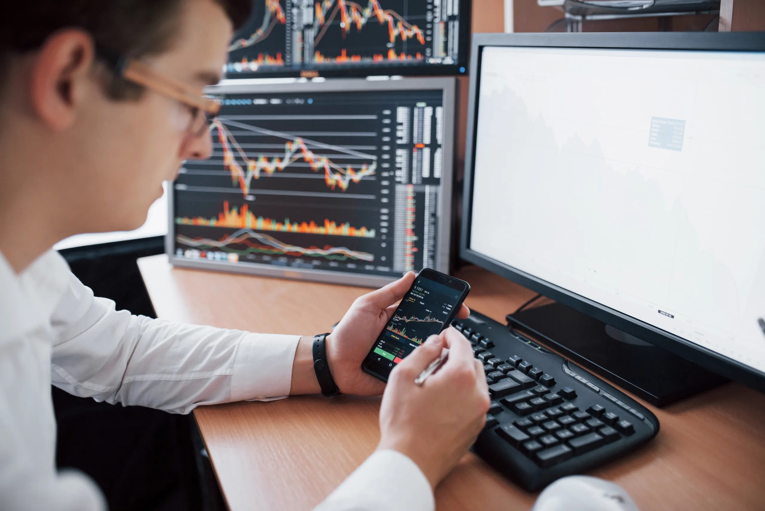 Over the shoulder view of and stock broker trading online while accepting orders by phone. Multiple computer screens ful of charts and data analyses in background.