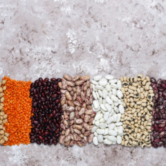 Imagem mostra diferentes tipos de feij&otilde;es e leguminosas alinhados lado a lado sobre um fundo texturizado, destacando variedade e cores do gr&atilde;o brasileiro.