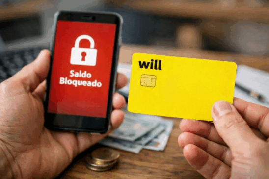 Imagem horizontal realista mostrando uma mão segurando um cartão do Will Bank diante de um smartphone com mensagem de saldo bloqueado na tela. A cena representa clientes enfrentando dificuldades para acessar valores na conta digital, ilustrando reportagens sobre instabilidade ou bloqueio de saldo no Will Bank. Composição limpa, iluminação natural e elementos financeiros ao fundo reforçam o contexto bancário.