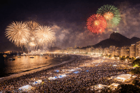 Imagem aérea e realista do Réveillon no Brasil, com destaque para a queima de fogos na praia de Copacabana, no Rio de Janeiro. Milhares de pessoas ocupam a orla iluminada, enquanto fogos de artifício coloridos explodem sobre o mar e o skyline urbano, com prédios residenciais e pontos turísticos ao fundo. A cena simboliza o impacto econômico do Réveillon na economia brasileira, refletindo o aumento do turismo, consumo e movimentação de serviços durante a virada do ano.