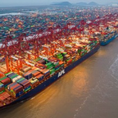 Imagem horizontal e realista de um grande porto internacional de importacao, com cont&ecirc;ineres mar&iacute;timos empilhados e guindastes em opera&ccedil;&atilde;o. &Agrave; esquerda, h&aacute; maior volume de cargas em tons de vermelho, remetendo &agrave; China e ao crescimento das exporta&ccedil;&otilde;es chinesas. &Agrave; direita, cont&ecirc;ineres em tons de azul e cinza representam os Estados Unidos, com menor movimenta&ccedil;&atilde;o portu&aacute;ria, simbolizando a retra&ccedil;&atilde;o no com&eacute;rcio externo. Ao fundo, um mapa-m&uacute;ndi discreto destaca China e EUA, refor&ccedil;ando o contexto de importacao e com&eacute;rcio global entre as duas maiores economias do mundo. A cena transmite an&aacute;lise econ&ocirc;mica, fluxo de mercadorias e disputas no mercado internacional.