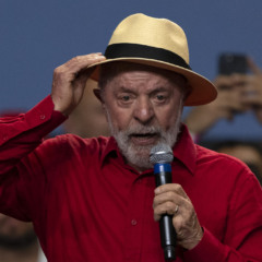 Imagem mostra LULA, presidente do Brasil, discursando em evento pol&iacute;tico com microfone na m&atilde;o. LULA veste camisa vermelha e chap&eacute;u claro, enquanto se dirige ao p&uacute;blico durante encontro com movimentos sociais. A cena ocorre em ambiente interno, com apoiadores ao fundo registrando o momento. A imagem representa a atua&ccedil;&atilde;o de LULA em agendas pol&iacute;ticas ligadas &agrave; reforma agr&aacute;ria, movimentos sociais e articula&ccedil;&atilde;o do governo federal em 2026.
