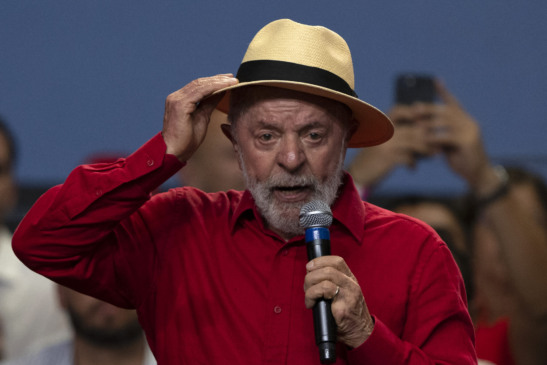 Imagem mostra LULA, presidente do Brasil, discursando em evento político com microfone na mão. LULA veste camisa vermelha e chapéu claro, enquanto se dirige ao público durante encontro com movimentos sociais. A cena ocorre em ambiente interno, com apoiadores ao fundo registrando o momento. A imagem representa a atuação de LULA em agendas políticas ligadas à reforma agrária, movimentos sociais e articulação do governo federal em 2026.
